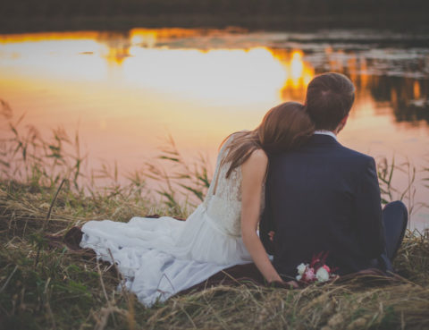 Photo of newlyweds sitting on bank of pond at sunset